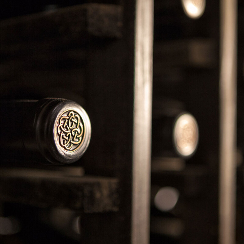 centennial mountain vineyard Close-up of a wine rack holding bottles, one cork displaying an intricate Celtic knot design, dimly lit setting.